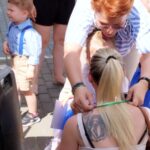 1716985030966 Na obrazku blondynkę z tatuażem na plecach, klęczącą, podczas gdy kobieta zawiera dodatkową wstążkę wokół jej ciała. Obok nich dziecko w niebieskim ubraniu na utwardzonej powierzchni.