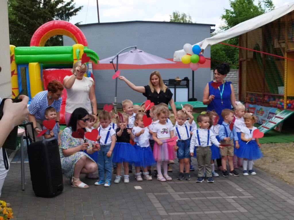 Na obrazku jest grupa dzieci ubranych w odświętne stroje, z ograniczeniami trzymającymi czerwone papierowe serduszka, pozujących do zdjęć wraz z dorosłymi na świeżym powietrzu. W tle znajdują się dekoracje, w tym dmuchanym domku do zabawy i balonów.