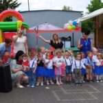 1716985031002 Na obrazku jest grupa dzieci ubranych w odświętne stroje, z ograniczeniami trzymającymi czerwone papierowe serduszka, pozujących do zdjęć wraz z dorosłymi na świeżym powietrzu. W tle znajdują się dekoracje, w tym dmuchanym domku do zabawy i balonów.