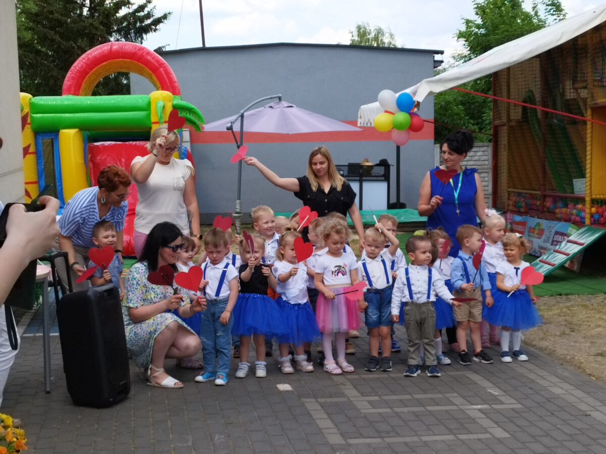 Na obrazku jest grupa dzieci ubranych w odświętne stroje, z ograniczeniami trzymającymi czerwone papierowe serduszka, pozujących do zdjęć wraz z dorosłymi na świeżym powietrzu. W tle znajdują się dekoracje, w tym dmuchanym domku do zabawy i balonów.