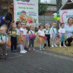 1716987301287 Na obrazku pojawia się grupa małych dzieci ubranych w kolorowe stroje, stojących i klęczących na otwartej, wybrukowanej przestrzeni. Towarzyszą im trzy osoby dorosłe, a w tle są baner i balony, co jest zawarte w nich lub w nich zawarte.