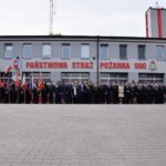 436401680_425130967131808_2309798235462292874_n Na obrazku występuje duża grupa ludzi w mundurach i formalnych strojach, stojących przed remizą strażacką, obok której powiewają różne flagi. Budynek ma znak wskazujący na Państwową Straż Pożarną i numer alarmowy 998.