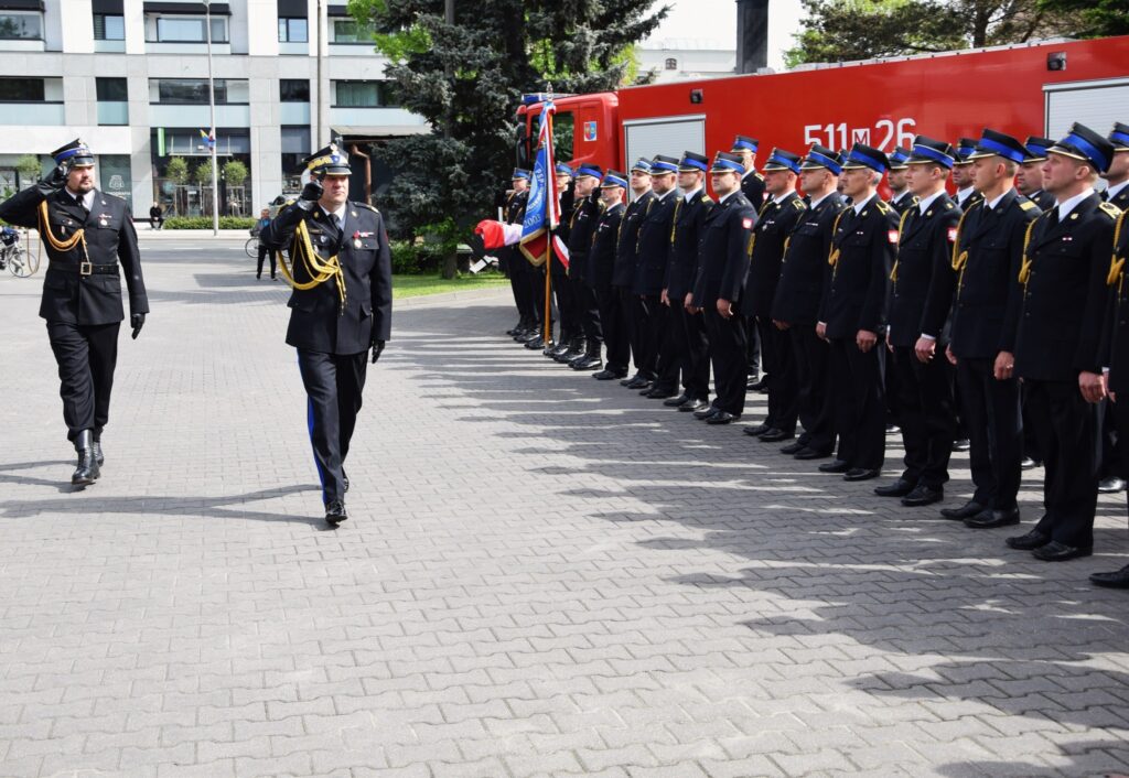 Dwóch umundurowanych mężczyzn salutuje sobie samodzielnie, podczas gdy grupa umundurowanych osób stoi w oczekiwaniu obok pojazdu strażackiego. Cała scena wygląda na formalną ceremonię lub podlega działaniu z pracą służb ratunkowych.