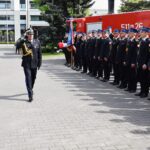 441163066_425130413798530_2010851356605956450_n Dwóch umundurowanych mężczyzn salutuje sobie samodzielnie, podczas gdy grupa umundurowanych osób stoi w oczekiwaniu obok pojazdu strażackiego. Cała scena wygląda na formalną ceremonię lub podlega działaniu z pracą służb ratunkowych.