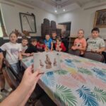 Na obrazku widocznym grupy dzieci stojących wokół stołu z kolorowym obrusem. Na pierwszym planie trzyma broszurę, na której znajdują się zdjęcia.