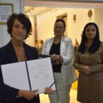 Na zdjęciu trzy kobiety w ramionach, z których jeden trzyma dokument z pieczęcią. Stoją jeden przed półką, której są książki i nagrody.