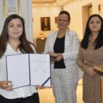 Na zdjęciu trzy kobiety w galerii, z których jedna trzyma certyfikaty nagrody. Pozostałe dwie kobiety oklaskują jej funkcję.