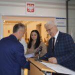 Na obrazku ceremonię z dwóch mężczyzn i jednej kobiety. Jeden z mężczyzn podpisuje dokument na podium z mikrofonem w pomieszczeniu, gdzie jest polska flaga.