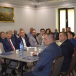 Na zdjęciu grupa dorosłych, która siedzi w sali konferencyjnej i słucha prezentacji. Inne z nich zwróceni są w stronę kamer.