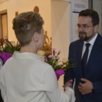 Na obrazku widoczny jest mężczyzna w garniturze, który uścisza dłoń inna osoba ubrana w kremowym stroju, w tle przypominającym galerię. Obok nich kobieta przechowująca bukiet kwiatowy.