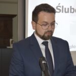 Na obrazku mężczyzn w niebieskim garniturze i okularach, który przemawia do mikrofonu za mównicą. W tle widać ekran prezentacyjny.