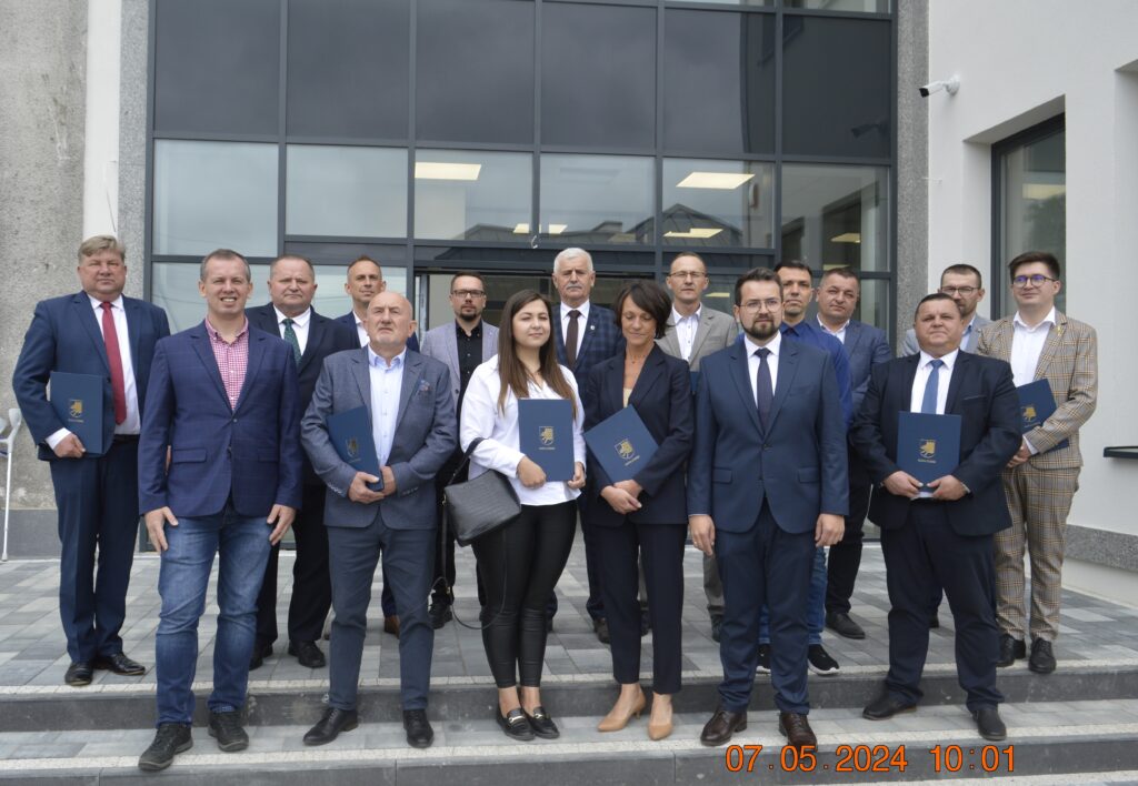 Na zdjęciu posegregowana grupa czternastu konkurencyjnych firm trzymających certyfikaty. Stoją oni na tle budynku i pozują do zdjęć.