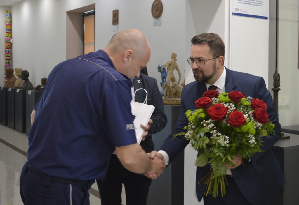 Na obrazku mężczyzny w garniturze i okularach ściskanych w dłoni mężczyzn w mundurze, trzymanych przy tym bukietu czerwono-białych kwiatów. Znalezienie się w pomieszczeniu, w którym znajduje się informacja, jest tablicą pamiątkową.