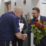 Na obrazku mężczyzny w garniturze i okularach ściskanych w dłoni mężczyzn w mundurze, trzymanych przy tym bukietu czerwono-białych kwiatów. Znalezienie się w pomieszczeniu, w którym znajduje się informacja, jest tablicą pamiątkową.