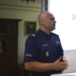 Na obrazku jest osoba w mundurze policyjnym, która stoi przed konsolą prezentacyjną i rozmówcą. W tle znajduje się drewniana szafka oraz mikrofon.