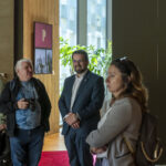 Na obrazku znajdują się trzy osoby, które są dobrze oświetlone, rozmawiają ze sobą. Jedna z nich trzyma urządzenie fotograficzne, a w tle jest duże okno oraz roślina.