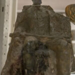 Na miejscu pojawia się brązowa rzeźba siedząca jako postać z zamyśloną miną, ustawiona na półce wystawowej, której widoczne są inne artefakty w tle. Otoczenie ograniczone przestrzenią muzealną lub galerią sztuki.