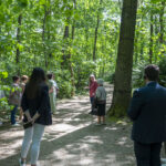 Na obrazku widać grupę osób stojących w zalesionym terenie, z których wydzielane są eleganckie. Ludzie ci są zaangażowani w działania i bacznie zauważają się w otoczeniu.