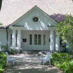 Na miejscu pojawia się biały dom z dwuspadowym dachem, dostępny z drzewami i zielenią. Na ścieżce do wejścia są dwie białe ławki.