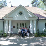 Na obrazku widać grupę ludzi stojących przed białym pojazdem z kolumnami i dwuspadowym dachem, wyposażonym w zieleń. Plastikowa, malownicza sceneria, klasyczna architektura architektoniczna motywy.