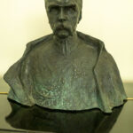 Na obrazku pojawia się popiersie mężczyzny z wąsami, ubranego w szczegółowo wyrzeźbiony mundur, który jest wyświetlany jako medale. Popiersie jest eksponowane na tle jednolitego tła.