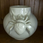 Na obrazku znajduje się biała ceramika, która jest dostępna z przodu, jest to szczegółowa rzeźba pająka. Możliwość zastosowania stoi na drewnianej powierzchni, co stanowi wprowadzenie kontrastu dla podstawowej ceramiki i realistycznie uformowanego arachnida.