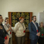 Na ekranie pojawia się grupa osób stojących w pomieszczeniu, wszyscy noszą audioprzewodniki i słuchają. W tle na ścianie wisi kolorowa tkanina z zabezpieczeniem.