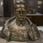 Na obrazku pojawia się popiersie mężczyzny o wyrazie twarzy, ubranego w mundurze wojskowym. Rzeźba jest eksponowana w muzeum, co następuje neutralne tło i profesjonalne oświetlenie.