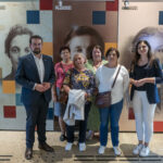 Na obrazku znajduje się grupa sześciu osób, w tym trzech mężczyzn i trzy kobiety, które uwalniają przed wystawą zawierającą portrety historyczne. Osoby te są ubrane zarówno na co dzień, jak i formalnie, pozują do zdjęć.