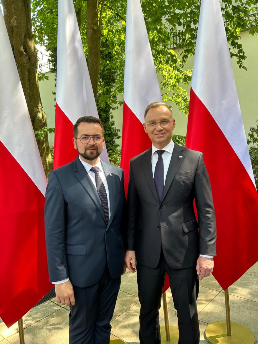 Na obrazku widać dwóch mężczyzn w garniturach, które są udostępniane przed uruchomieniem flagami polskimi i reagują na aparat. W tle można znaleźć drzewo i bujną roślinność.