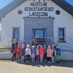 Na wolności jest grupa dzieci oraz dorosła osoba przed wejściem do „Muzeum Konstantego Laszczyka”. Tło plastikowe przezroczyste, błękitne niebo.