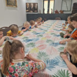 Na obrazku dzieci siedzących przy stole, na którym znajduje się symbol obrusu przedstawiony na liściastym produkcie. Dzieci uczestniczą w twórczej zabawie z gliną, tworząc formę kształtową i formy.