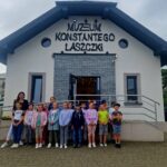 Na obrazku grupy dzieci oraz jednego dorosłego, którzy pozują do zdjęć przed wbudowanym z szyldem "Muzeum Konstantiego Laszczki". W tle jest fasada muzeum poświęconego twórczości rzeźbiarza Konstantina Laszczki.