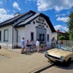 Na obecność grupy ludzi, w tym dzieci z rowerami, należy przed Muzeum Konstantego Laszczki. Na pierwszym planie jest beżowy kabriolet, całość rozgrywająca się w słonecznym dniu z błękitnym niebem i chmurami.