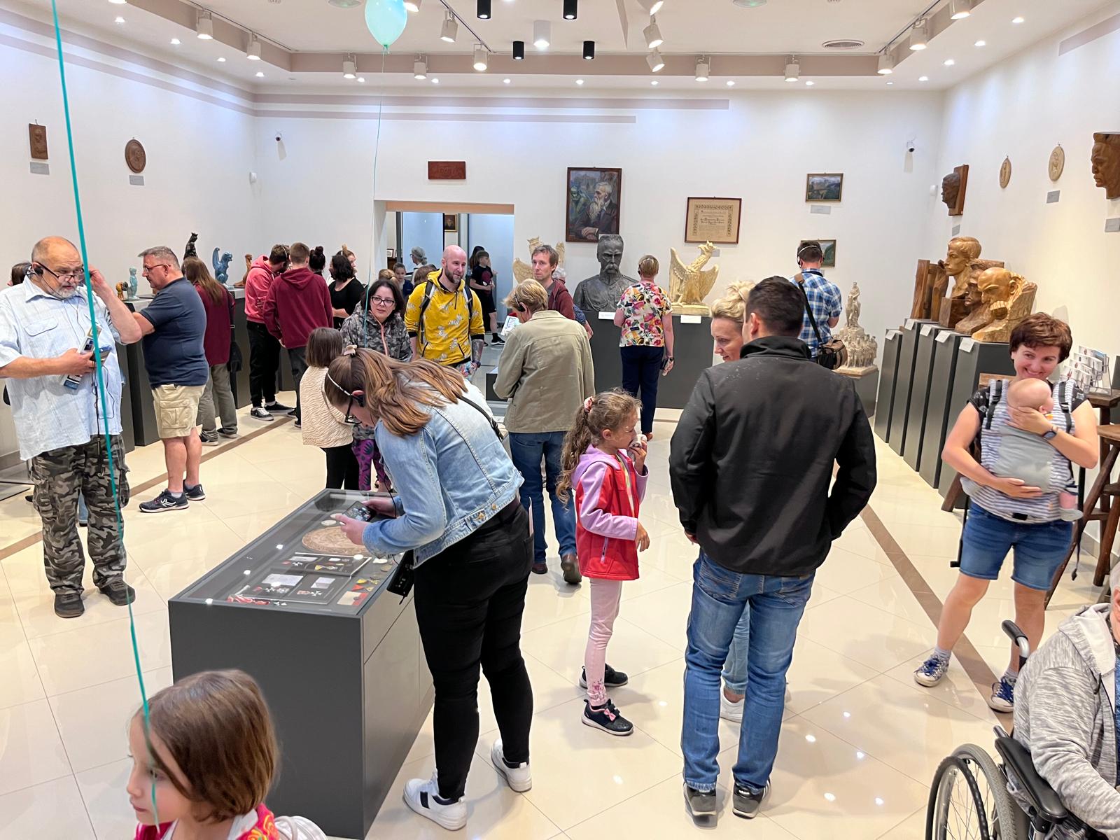 Na obrazku jest grupa osób, w tym dzieci, które zwiedzają wystawę muzealną. Osoby obserwowane przez inspektorów szklanych gablotach, podczas gdy inni przechodzą wokół lub siedzą; jest dobrze oświetlone z artefaktami na ścianach i postumentach.