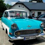 Na obrazku pojawia się klasyczny samochód marki Opel w cechach turkusowych i białej z polską tablicą rejestracyjną, ujawnioną przed domem. W miejscu dostępnym jest osoba znajdująca się obok innego samochodu.