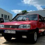 Na zewnątrz znajduje się czerwony samochód z tablicą rejestracyjną „KWI 05TE”, wprowadzony na zewnątrz w słoneczny dzień. W tle widoczne są kilka budynków oraz inny pojazd.