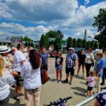 Na obrazku widać grupę ludzi zgromadzonych na świeżym powietrzu w słoneczny dzień, stojących na wybrukowanej przestrzeni w pobliżu budynków i zieleni. Część osób rozmawia ze sobą, gdy inni patrzą lub trzymają rowery.