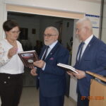 Na obrazku znajdują się trzy osoby ubrane w formalnych ubraniach, które są częścią wnętrza budynku. Kobieta po lewej stronie trzyma plakietkę, a dwóch mężczyzn obok niej wygląda na to, że jest ona bezpośrednio trzymana i jednocześnie trzyma ją. Na zdjęciu widnieje również stempel czasowy z inspektorem „27.03.2024 14:23”.