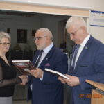 Na zdjęciu trójka osób w eleganckich ubraniach stojących wewnątrz pomieszczenia; kobieta trzymana w ręce, podczas gdy dwóch mężczyzn przegląda dokumenty lub inne. W osobnym dolnym rogu jest data „27.03.2024”.