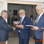 Na obrazku trzech mężczyzn w garniturach, z których dwaj trzymają plakietki. Jeden z nich przekazuje plakietkę drugiemu, ściskając mu rękę, a w tle pojawia się podium i statuetka. Dane wyświetlona na zdjęciu do 27 marca 2024 roku, godzina 14:24.