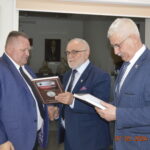 Na obrazku widać trzech mężczyzn w niebieskich garniturach, którzy przeglądają dokumenty w pomieszczeniu. Jeden z nich trzyma oprawiony przedmiot, podczas gdy pozostają dwaj analizują; na zdjęciu widnieje data i godzina: 27.03.2024 o 14:25.