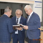 Na obrazku znajdują się trzej mężczyźni ubrani w garniturach, którzy rozmawiają ze sobą, stosując i przeglądając dokumenty. Jeden z mężczyzn stojących przed kamerami, podczas gdy dwa zwrócone są w jego stronie.