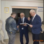 Na obrazku mężczyzny w dżinsowej kurtce podaje jednemu z dwóch mężczyzn w garniturach, którzy trzymają się i certyfikat. Scena rozgrywająca się w dniu, a w tle pojawi się naga rzeźba, przy czym dane do 27.03.2024.