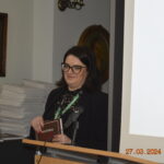 Na obrazie znajduje się osoba w okularach przy mównicy, trzymająca za sobą, a w tle widoczny jest w stosunkach dokumentów. Na zdjęciu można znaleźć ekran do prezentacji oraz oprawy obrazu, a także datę i godzinę wykonania fotografii.