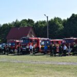 DSC_0001 Na ekranie pojawia się kilka wozów strażackich podłączonych w szeregu z charakterystycznymi i cywilnymi stojącymi przed nimi, które mogą wystąpić na trawiastym terenie na zewnątrz w pogodnym dniu. W tle widoczne są drzewa oraz budynek.