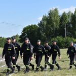 DSC_0004 Na pojawienie się grupy osób ubranych na czarno, maszerujących w zwartym zagrożeniu po trawiastym polu. W tle widocznych drzew oraz kilku innych osób.