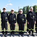 DSC_0021 Na obrazie widać grupę młodych osób w czarnych mundurach i beżowych czapkach, które są uwalniane w szeregach na świeżym powietrzu w słoneczny dzień. W tle widać drzewa i roślinność.