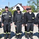 DSC_0026 Na obrazku pojawia się grupa sześciu osób ubranych w czarne mundury strażackie z naszymiywkami „STRAŻ”, stojącymi w szeregu na świeżym powietrzu. Część z nich ma na głowach czapki, a za nimi widoczne są okna mieszkalne.