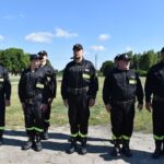 DSC_0029 Na zewnątrz występuje grupa osób w mundurach stojących w szeregu na zewnątrz podczas promieni słonecznych dnia. Mają oni na sobie czarne mundury z neonowymi, żółtymi paskami przy sekcji części spodniej oraz czapki.